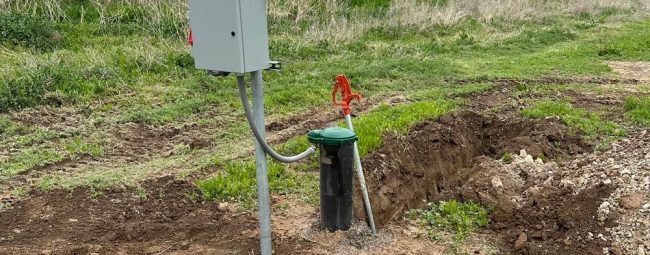 A water pump station with a control box and a green-handled valve is installed outdoors, surrounded by grassy fields and hills.