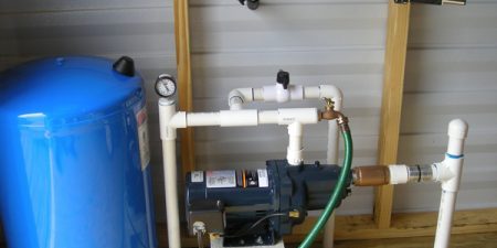 A blue water storage tank is connected to a water pump via pipes and fittings, installed in a room with wooden framing.