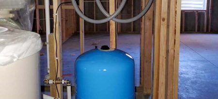 A blue water storage tank is connected to pipes and valves in an unfinished room, part of a residential water pumping system.