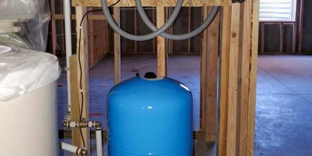 A blue water storage tank is connected to pipes and valves in an unfinished room, part of a residential water pumping system.